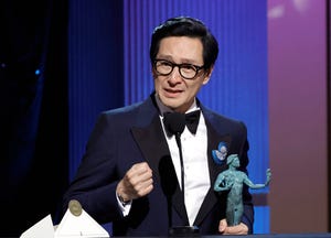 ke huy quan, wearing a black tuxedo, stands in front of a microphone on a stage, holding a sag award trophy in his left hand and clinching his right fist