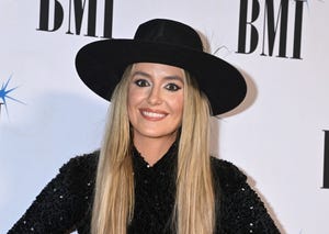 lainey wilson smiles at camera, she wears a black wide brimmed hat with a black sequin top