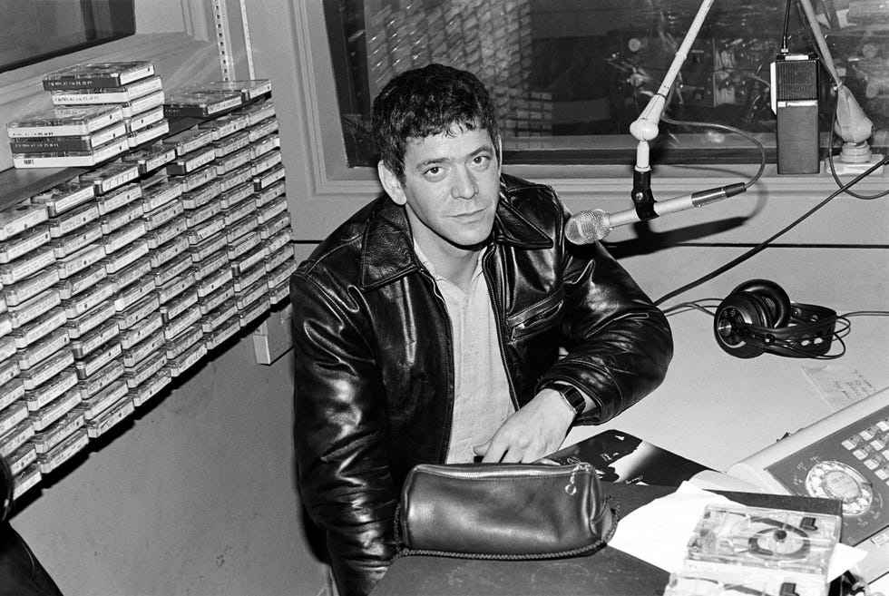 lou reed sits at a table with a microphone hanging in front of him, he looks at the camera and wears a leather jacket and shirt