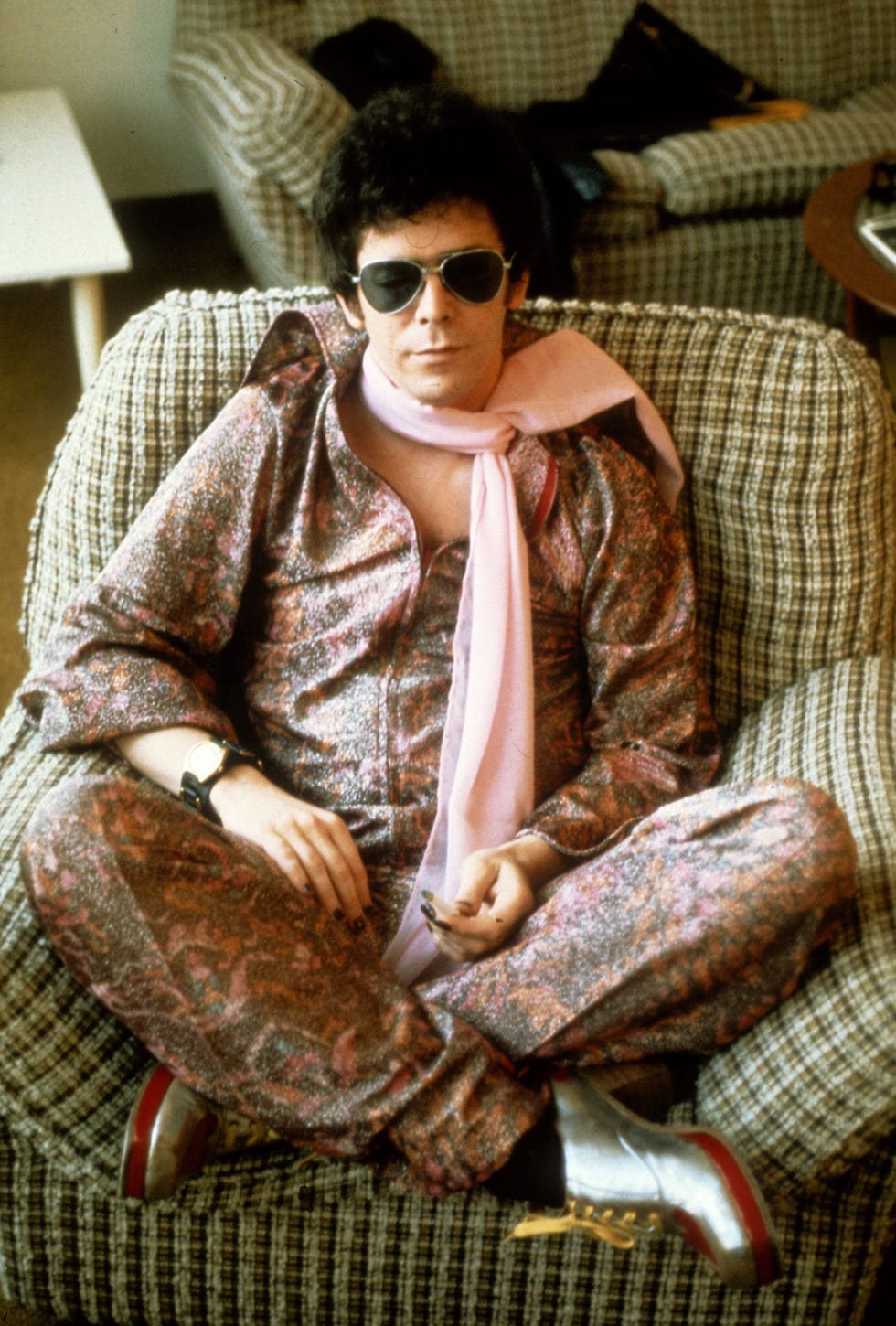 lou reed sits crosslegged in an armchair wearing a pink patterned onesie, pink scarf and aviator sunglasses