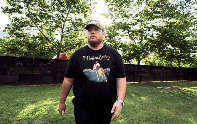 luke combs walks in a grass field, he looks to the left and wears a black graphic t shirt and pants with a baseball cap, sunlight filters through trees behind him