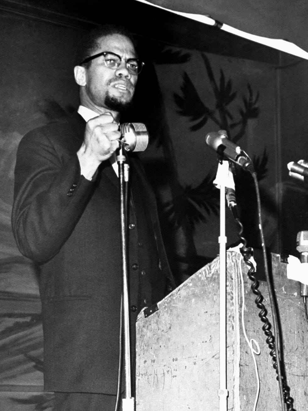 malcolm x speaks into microphones while standing behind a podium, he wears a suit jacket, collared shirt, and glasses