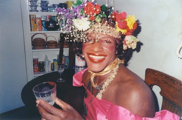 marsha p johnson smiles at the camera while seated at a table, she wears a flower crown and a bright pink dress and holds a glass