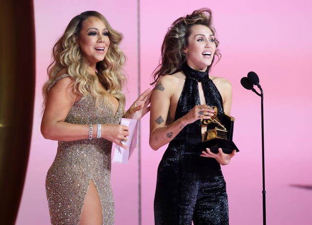 miley cyrus holding a grammophone trophy and smiling while mariah carey congratulates her from behind