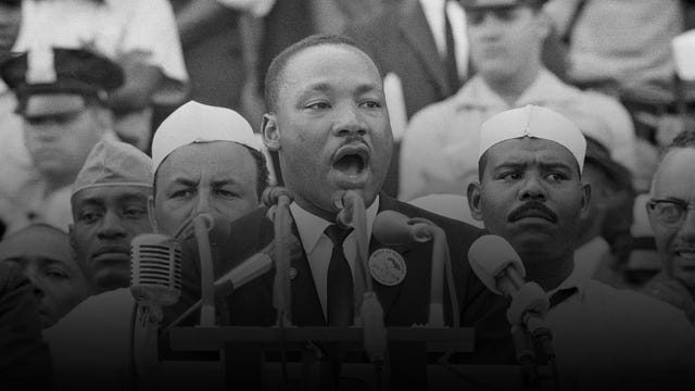 martin luther king jr speaks into several microphones as men stand behind him, he wears a suit jacket, collared shirt and tie with a button on his lapel