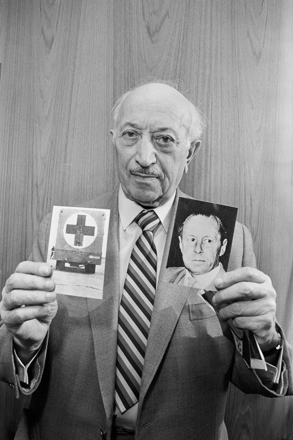 simon wiesenthal looks at the camera and holds up a photo in each hand, one shows a medical vehicle, the other shows a man looking to the right