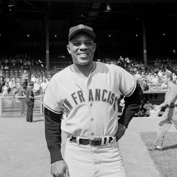 willie mays stands on a baseball diamond and smiles, he wears a baseball hat and uniform for the san francisco giants