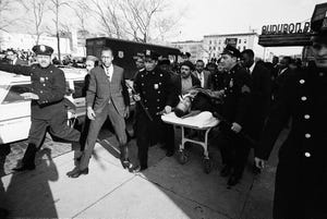 police roll a stretcher with malcolm x body on a sidewalk, many people are on the street as well as a police truck and cars