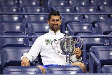 novak djokovic sits in a stadium seat and smiles at the camera, he holds a large silver trophy that rests on his leg and wears a white jacket with the number 24 on it and blue shorts