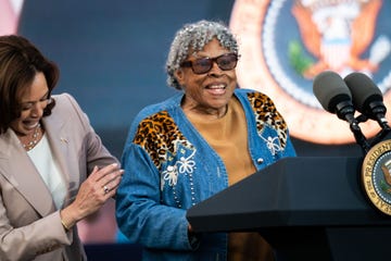 opal lee, wearing a denim designed coat, yellow shirt, and sunglasses, speaks at a podium with two microphones and the us presidential seal, standing next to a laughing kamala harris