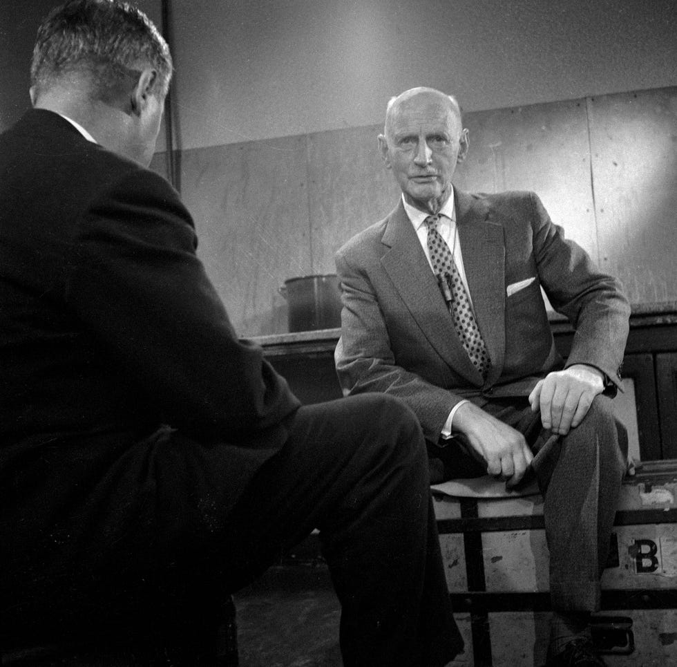 otto frank looks at the camera while seated, he wears a suit and dress shirt with a patterned tie