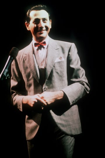 paul reubens as pee wee herman on a darkened stage, standing in front of a microphone and smiling