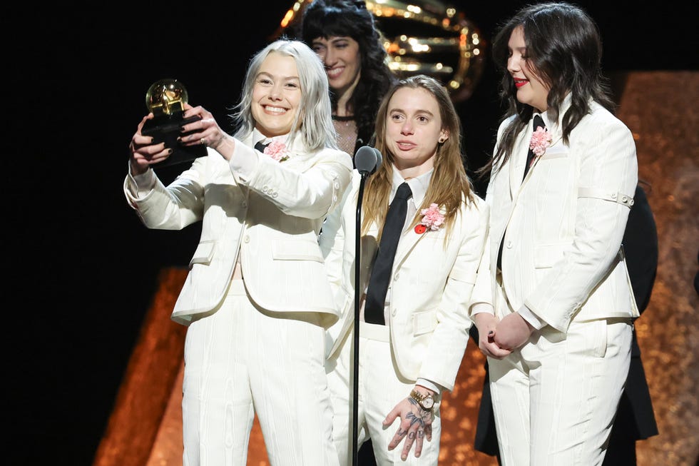 phoebe bridgers smiles and holds up a grammy trophy while standing next to julien baker and lucy dacus, all three women wear matching white suits with black ties and pink flower boutonnieres
