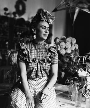 frida kahlo sits on a table while wearing a floral head piece, large earrings, a plaid blouse and striped pants, she looks off to the right