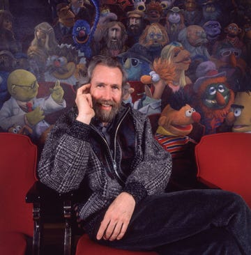 jim henson smiles at the camera as he holds one hand up to his face and sits in a red theater seat, behind him is a mural of many muppets, he wears a gray zip up jacket and dark pants