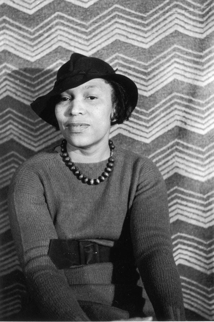 zora neale hurston wearing a sweater and hat and sitting for a photo in front of a striped backdrop