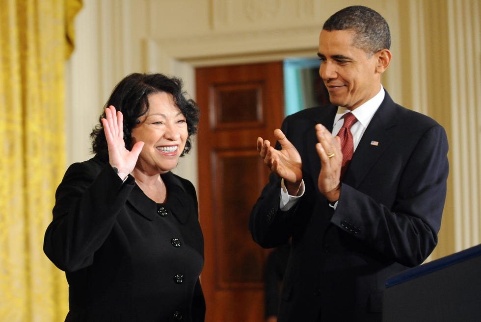 sonia sotomayor smiles and waves to an off camera crowd, earing a black dress, while barack obama smiles and applauds next to her, wearing a black suit and red tie