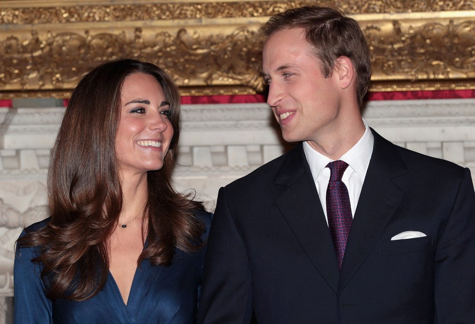 kate middleton and prince william look at each other and smile, she wears a blue dress, he wears a black suit jacket with a white collared shirt and red and navy tie