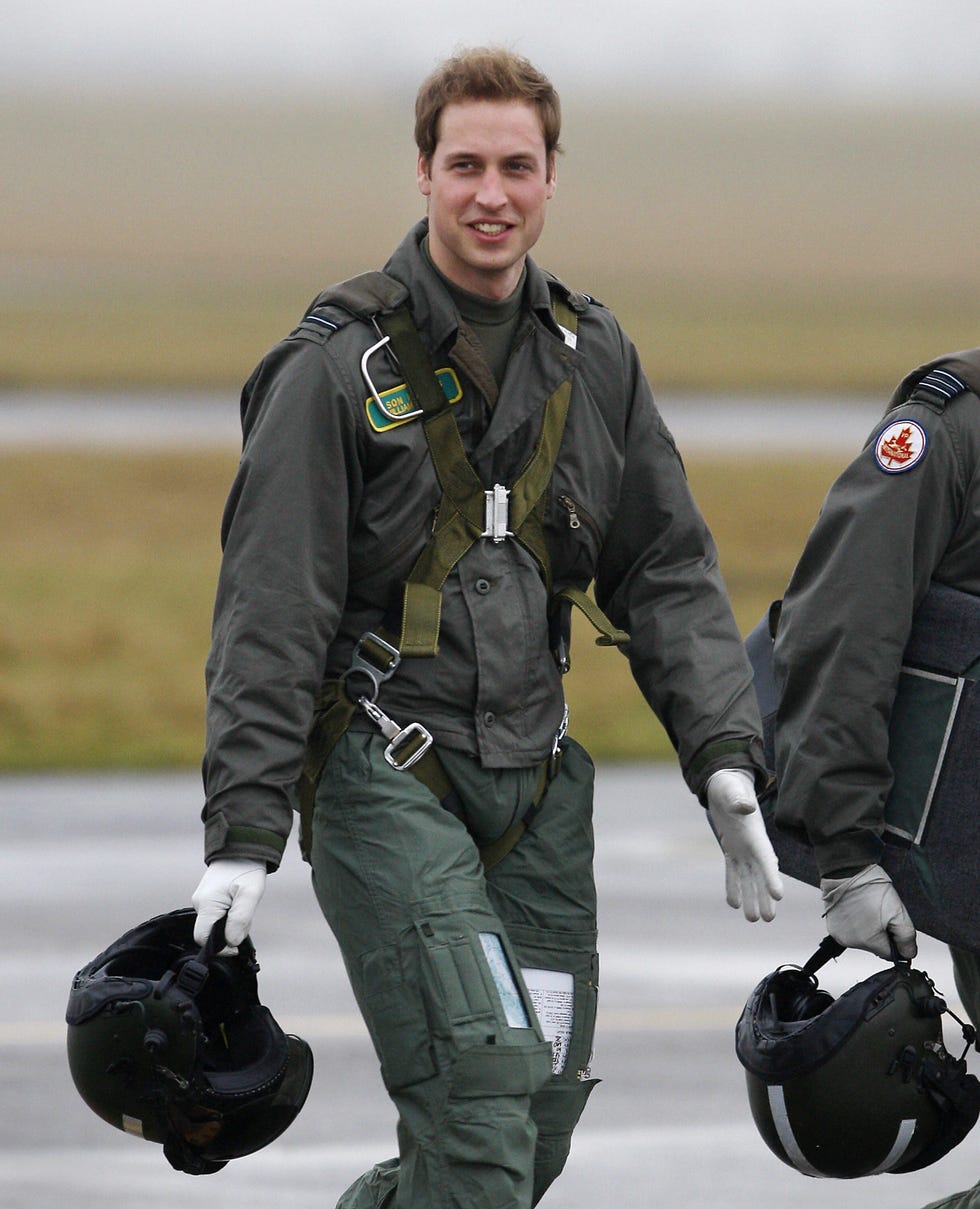 prince william walks across tarmac outside while wearing military fatigues, a harness and white gloves, he holds a helmet in one hand