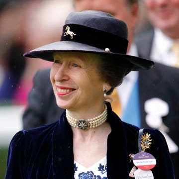 princess anne smiles and looks left, she wears a hat and blue velvet jacket over a white and blue top with brooches and pins, she also has on a pearl necklace and earrings