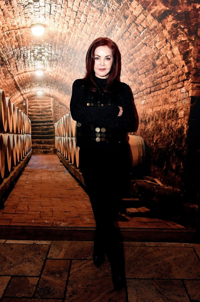 priscilla presley stands in front of several wine barrels in a stone arched cellar, she looks at a camera with a slight smile, her arms are crossed and she is wearing a black outfit