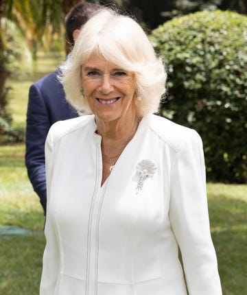 queen camilla smiles at the camera, she wears a white blouse with a pearl and diamond broach along with pearl earrings and simple gold necklaces