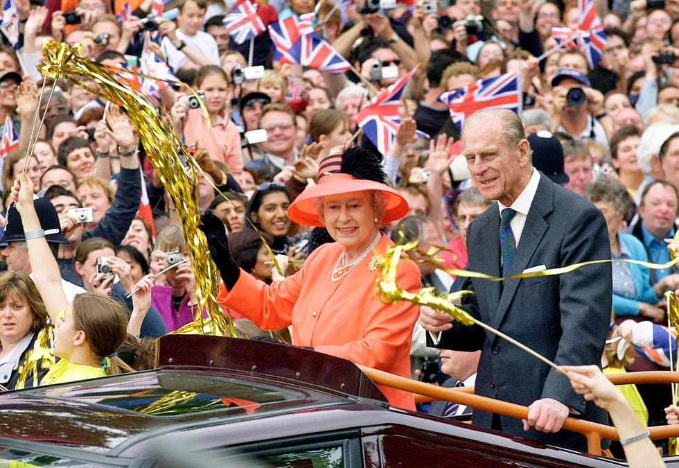 queen elizabeth ii and prince philip stand in the bed of a car that travels through crowds, both smile and wave as people wave british flags and golden streamers, the queen wears an orange outfit and matching hat, the prince wears a gray suit