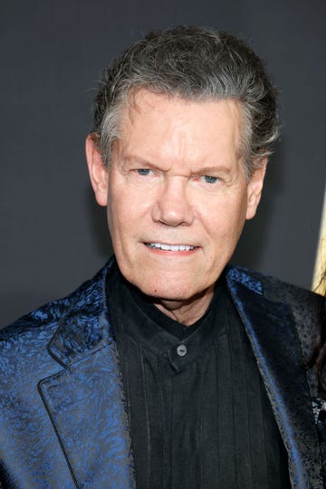 randy travis smiles at the camera, he wears a black shirt and blue and black patterned suit jacket