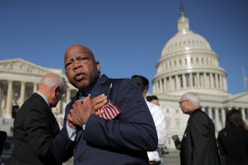congressman john lewis holds a small american flag to his chest, he stands outside the us capitol building in a suit jacket, other people stand in the background