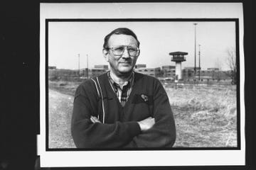 lionel dahmer stands outside a fenced complex with his arms crossed, he wears a sweater, plaid shirt, and large glasses