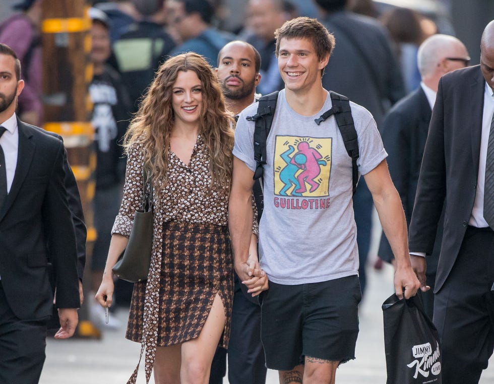 riley keough and ben smith petersen smile and walk hand in hand on a sidewalk, she wears a patterned button up shirt with a houndstooth pencil skirt, he wears a gray graphic t shirt and a backpack