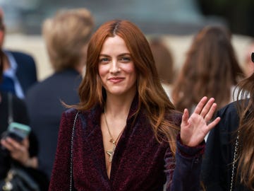 riley keough smiles and waves at the camera, she wears a burgundy jacket and golden star necklace