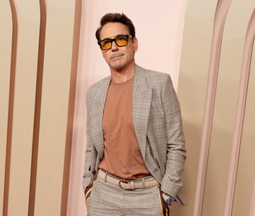 robert downey jr stands in a plaid suit with his hands in his pants pockets, he also wears orange tinted glasses with black frames and a rust orange tshirt