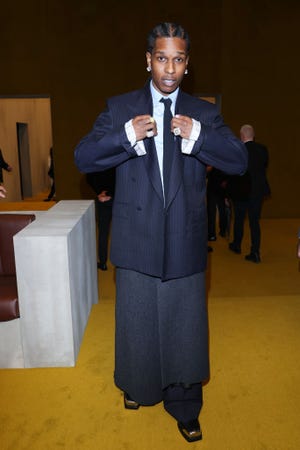 asap rocky looks at the camera as he adjusts his navy pinstripe suit jacket, he also wears a matching tie, white collared shirt, earrings and rings