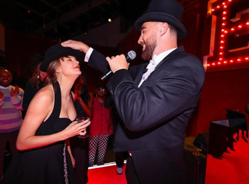 a celebration scene featuring a performer engaging with a guest both wearing hats