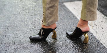 a pair of stylish heeled shoes with a black upper and shiny gold heels are shown on a rain soaked surface the footwear reveals a glimpse of the wearer's ankle, which is adorned with a small tattoo the surrounding area is slightly blurred, indicating a rainy environment, while the reflection of raindrops enhances the urban setting