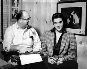 colonel tom parker looks at a smiling elvis presley while the men are seated inside a room, parker has a pipe in his mouth and a typewriter in front of him, pictures hang on the wall around them