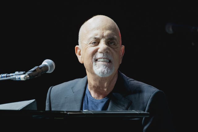 billy joel smiles at the camera as he sits at a piano, he wears a dark suit jacket and navy blue tshirt