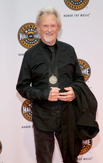 kris kristofferson smiles at the camera while standing in front of a white backdrop with country music hall of fame logos, he wears a black collared shirt and tie with a medallion hanging from his neck on a black strap