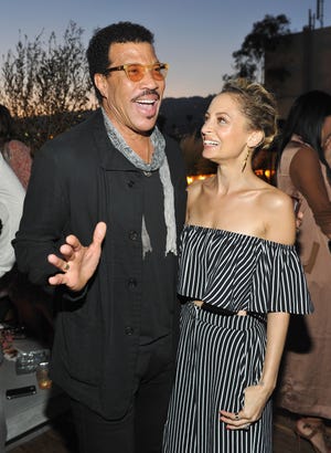 lionel richie and nicole richie stand together at an event, lionel is smiling as he speaks and has one arm around nicole who is looking at lionel and smiling