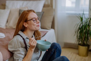 smiling woman with eyes closed eating food sitting in front of bed at home