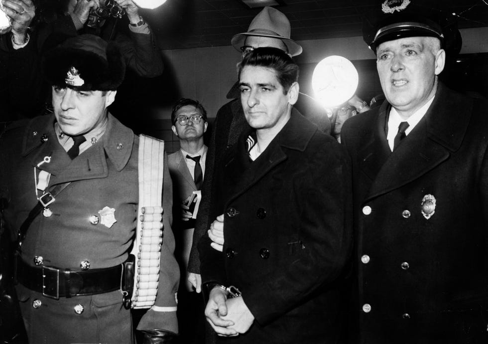 albert desalvo, wearing handcuffs and a dark peacoat, is led to prison by authorities, an officer is on either side of him, in the background are photographers, photography equipment and other men