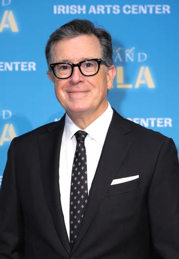 stephen colbert smiling for a red carpet photo in a black suit and patterned black tie