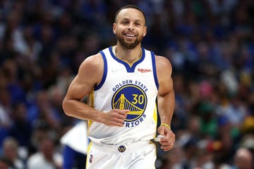 stephen curry smiles and looks past the camera, he wears a white golden state warriors jersey with blue and yellow accents