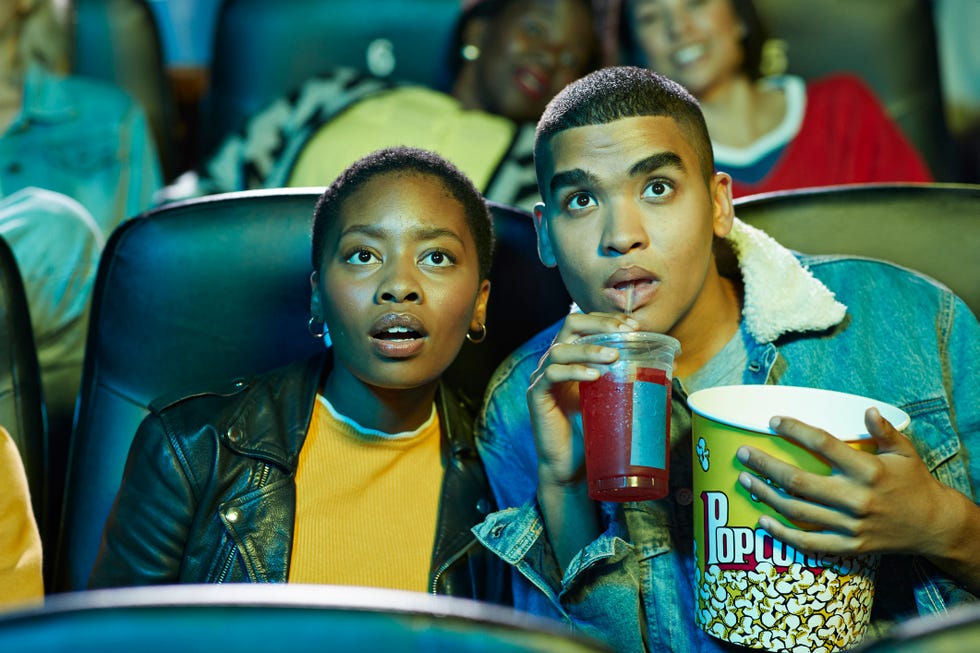 surprised young man drinking soda while watching movie with friend in cinema hall