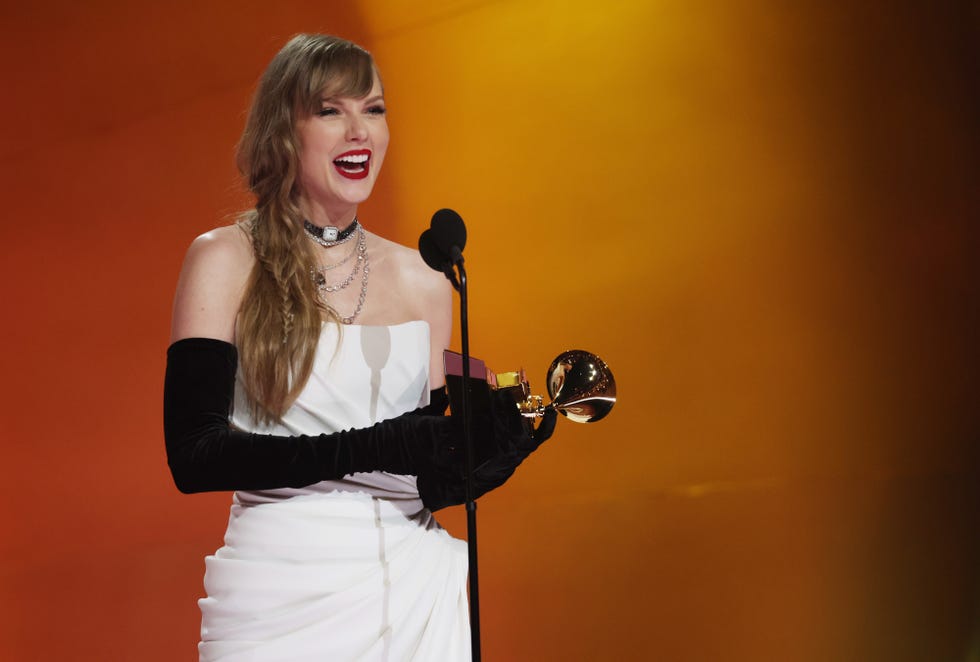taylor swift smiles as she stands at a microphone and holds a grammy trophy
