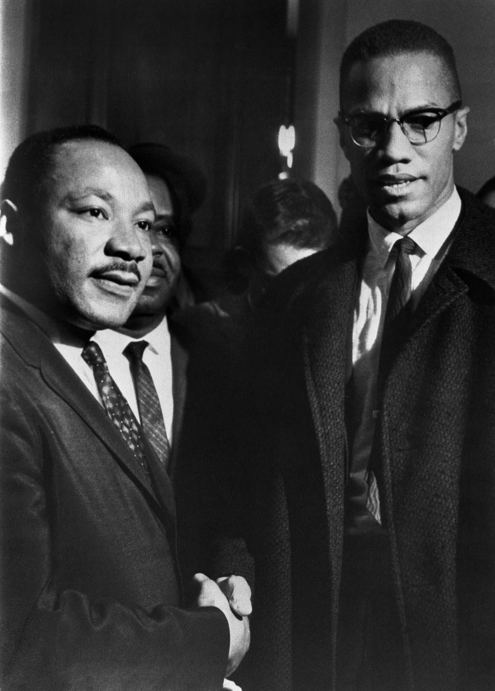 martin luther king jr and malcolm x shake hands while standing next to each other, both men wear collared shirts and ties with a suit jacket or coat