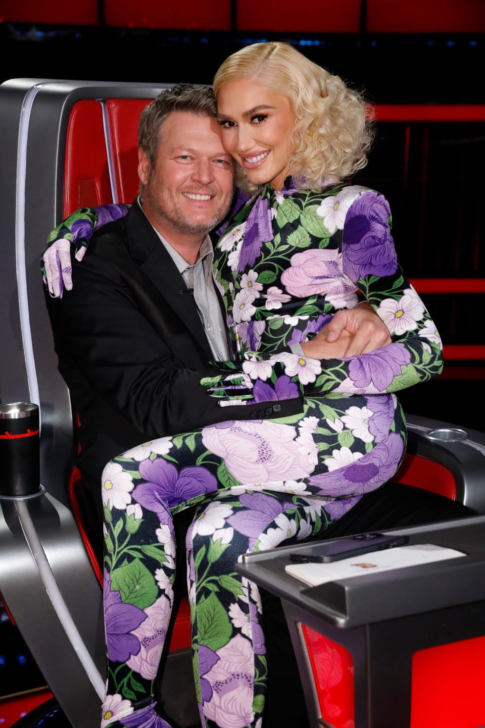 blake shelton sits in a red coach chair from the voice, gwen stefani sits on his lap, they hug each other and smile at the camera