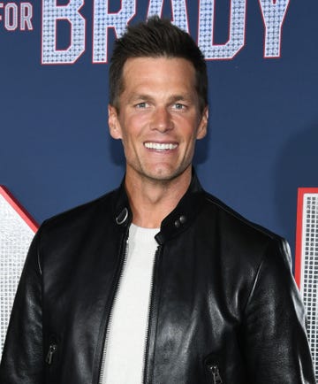tom brady smiles at the camera, he wears a black leather jacket over a white shirt and stands in front of a navy background