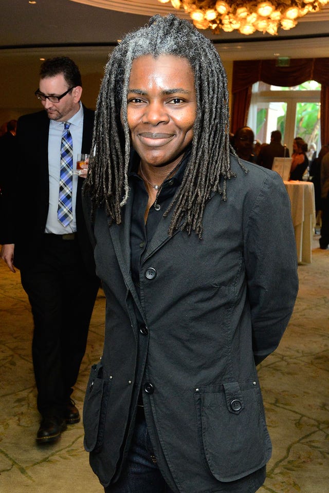 tracy chapman smiles at the camera while standing inside an event space with a chandelier, she wears a black jacket and black collared shirt, her dreads are slightly gray at the roots and reach past her shoulders